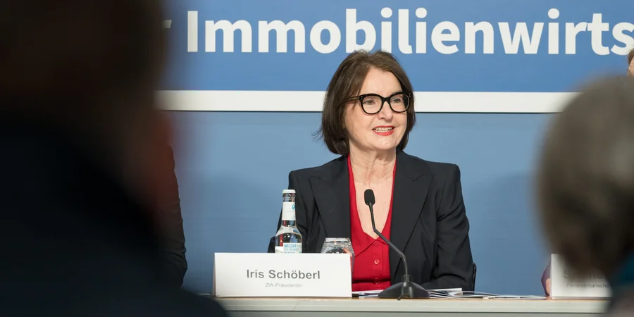 Fotografie einer Pressekonferenz mit Iris Schöberl, die an einem Podium vor blauem Hintergrund mit Aufschrift „Immobilienwirtschaft“ spricht. Namensschild, Mikrofon und Wasserflasche markieren den offiziellen Veranstaltungsrahmen; unscharfe Personen im Vordergrund zeigen die Perspektive des Publikums.