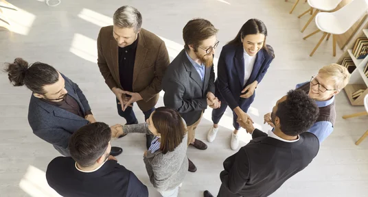 Gruppe von sieben Personen im beruflichen Kontext, die sich in einem hellen Büro zu einer Gesprächsrunde versammelt haben. Mehrere Personen reichen sich die Hand, während formelle Kleidung, offene Körperhaltung und kreisförmige Anordnung Zusammenarbeit und Teamkommunikation vermitteln.
