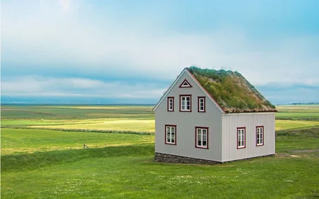 Fotografie eines alleinstehenden, weiß-grauen Hauses in weitläufiger grüner Küstenlandschaft. Das Haus besitzt rote Fensterrahmen, ein steiles Dach mit Grasbewuchs und steht vor flachem Ackerland unter bewölktem blauem Himmel.