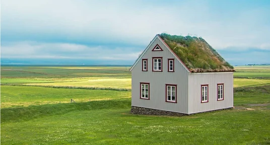 Fotografie eines alleinstehenden, weiß-grauen Hauses in weitläufiger grüner Küstenlandschaft. Das Haus besitzt rote Fensterrahmen, ein steiles Dach mit Grasbewuchs und steht vor flachem Ackerland unter bewölktem blauem Himmel.