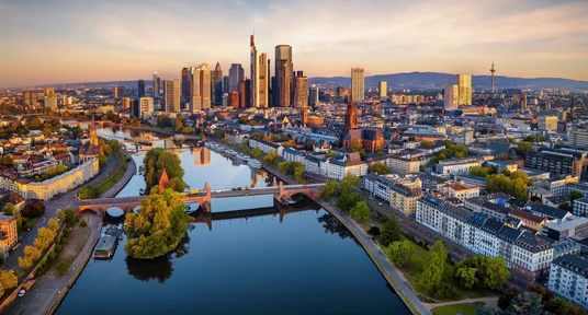 Luftaufnahme von Frankfurt am Main mit Skyline, Main und dicht bebautem Stadtgebiet im warmen Abendlicht. Im Vordergrund verbinden mehrere Brücken beide Flussufer, während markante Wolkenkratzer, darunter der Commerzbank Tower, die Stadtsilhouette prägen.