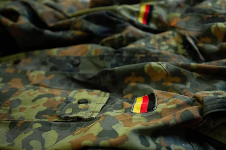 Fotografie mehrerer deutscher Bundeswehr-Tarnuniformen im Flecktarnmuster, vermutlich zur Darstellung militärischer Bekleidung und Ausrüstung. Sichtbar sind Taschen, Knöpfe und mehrere Aufnäher in Schwarz-Rot-Gold auf grün-braunem Stoff.