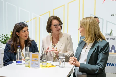 Fotografie von drei Frauen im Gespräch an einem Messestand oder Konferenztisch, vermutlich im Rahmen eines geschäftlichen Austauschs. Auf dem Tisch stehen Wasserflaschen, Gläser, Informationsmaterial und ein Tablet; gelbe Liniengrafiken sowie teilweise sichtbare Firmenlogos prägen den Hintergrund.