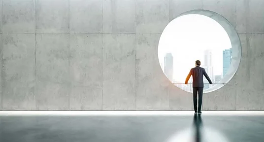 Fotografie eines Mannes in Anzug, der durch ein großes rundes Fenster auf eine Stadtlandschaft mit Hochhäusern blickt. Minimalistischer Raum mit glattem Betonwand und reflektierendem Boden betont Einsamkeit und Nachdenklichkeit.
