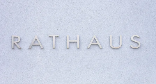 Fotografie eines hellen, strukturierten Gebäudeeingangs mit großem Schriftzug „RATHAUS“. Graue, dreidimensionale Buchstaben heben sich deutlich von hellblauer Wand ab und kennzeichnen ein Rathaus.