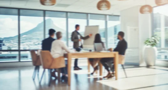 Fotografie einer modernen Bürositzung mit fünf Personen an einem Holztisch, vermutlich zur Präsentation oder gemeinsamen Besprechung. Eine stehende Person zeigt auf ein Flipchart, während große Fenster Berglandschaft zeigen; Hängelampen, Pflanzen und helle Einrichtung prägen den Raum.