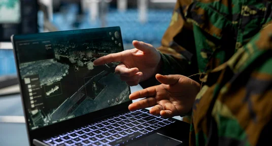 Fotografie zweier Personen in Tarnkleidung, die gemeinsam eine digitale Karten- oder Geodatenansicht auf einem Laptop auswerten. Eine Person zeigt auf Küstenlinien und markierte Bereiche am Bildschirm, während beleuchtete Tastatur und interaktive Kartenelemente technische Analyse und Einsatzplanung verdeutlichen.