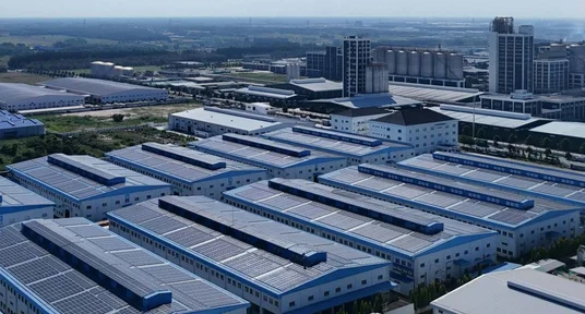 Luftaufnahme eines weitläufigen Industrieparks mit zahlreichen Produktions- und Lagerhallen. Großflächige Solarpaneele bedecken viele blau-weiße Dächer, während im Hintergrund Hochhäuser, Verkehrswege und flache Landschaft sichtbar sind.