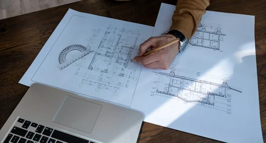 Fotografie eines Arbeitsplatzes mit architektonischen Bauplänen, auf denen eine Hand mit Bleistift Grundrisse und technische Details markiert. Ein Laptop, Winkelmesser und weitere Planunterlagen verweisen auf Planung oder Prüfung eines Bauprojekts.