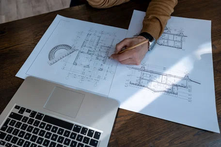 Fotografie eines Arbeitsplatzes mit architektonischen Bauplänen, auf denen eine Hand mit Bleistift Grundrisse und technische Details markiert. Ein Laptop, Winkelmesser und weitere Planunterlagen verweisen auf Planung oder Prüfung eines Bauprojekts.