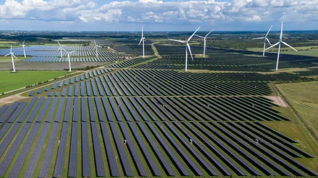 Luftaufnahme eines kombinierten Solar- und Windparks zur Darstellung erneuerbarer Energiegewinnung. Dicht angeordnete blaue Photovoltaikmodule bilden lange Reihen über grünen Feldern, ergänzt durch mehrere weiße Windräder unter bewölktem Himmel.