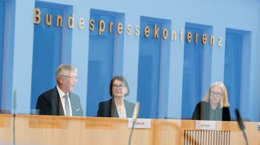 Fotografie einer Bundespressesekonferenz mit drei Personen an einem Podium und Mikrofonen. Blaue Glaswand mit goldener Aufschrift „Bundespressekonferenz“, Namensschilder auf dem Tisch und Spiegelungen im Vordergrund prägen die Szene.