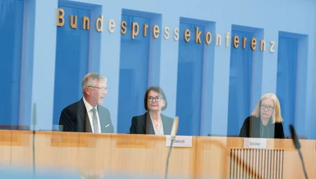 Fotografie einer Bundespressesekonferenz mit drei Personen an einem Podium und Mikrofonen. Blaue Glaswand mit goldener Aufschrift „Bundespressekonferenz“, Namensschilder auf dem Tisch und Spiegelungen im Vordergrund prägen die Szene.