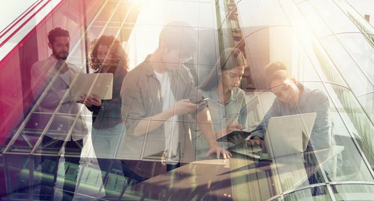 Fotografie einer vielfältigen Gruppe von Berufstätigen bei gemeinsamer Arbeit mit Laptop, Tablet und Unterlagen in modernem Glasbüro. Transparente Architektur, Sonnenlicht und Überblendungen vermitteln Zusammenarbeit, Digitalisierung und dynamische Arbeitsumgebung.