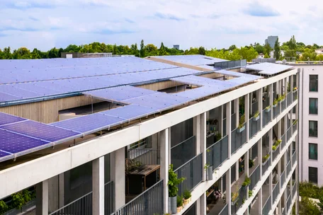 Luftaufnahme eines modernen Mehrfamilienhauses mit großflächiger Photovoltaikanlage auf dem Flachdach. Solarmodule bedecken mehrere Dachflächen, während Balkone, begrünte Bereiche und Bäume nachhaltige Architektur im urbanen Umfeld hervorheben.