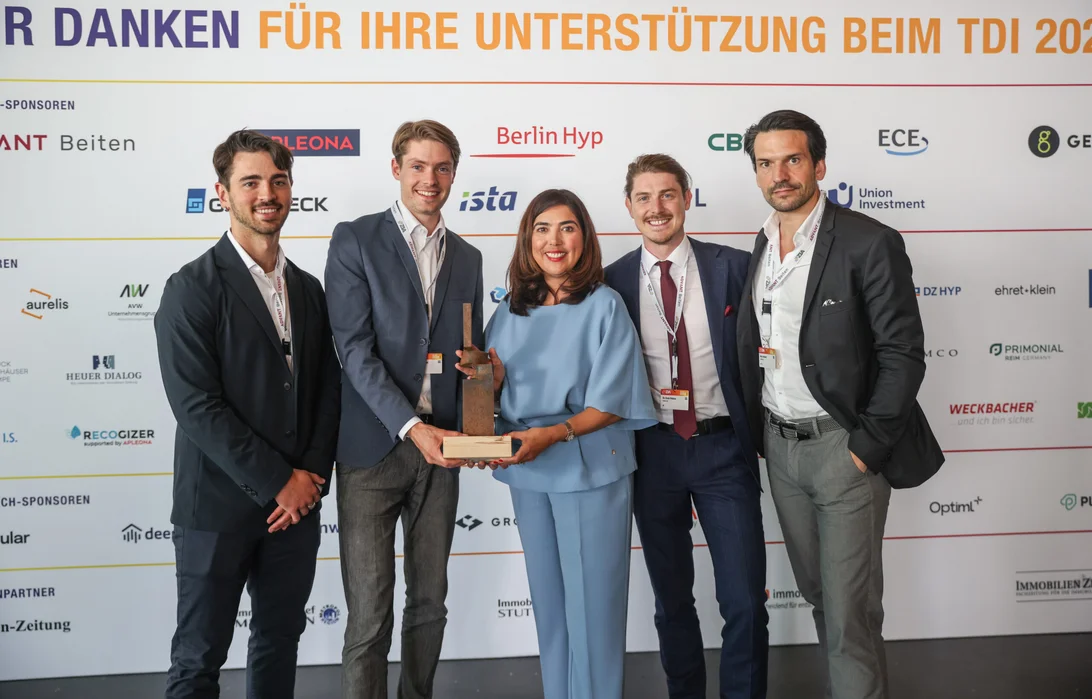 Fotografie von ZIA-Hauptgeschäftsführerin Aygül Özkan und den Gewinnern des PropTech of the Year Awards 2024, Optiml, die vor einer Sponsorenwand stehen und einen Pokal halten. Szene zeigt feierlichen Moment bei einer Veranstaltung, hervorgehoben durch Logos von Unternehmen und Organisationen im Hintergrund.