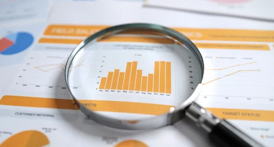 Fotografie einer Lupe auf Geschäftsberichten zur Analyse von Unternehmenskennzahlen. Orange Balkendiagramme, Kreisdiagramme und Liniengrafiken zeigen Umsatz-, Kunden- und Statusdaten mit monatlichen Trends.