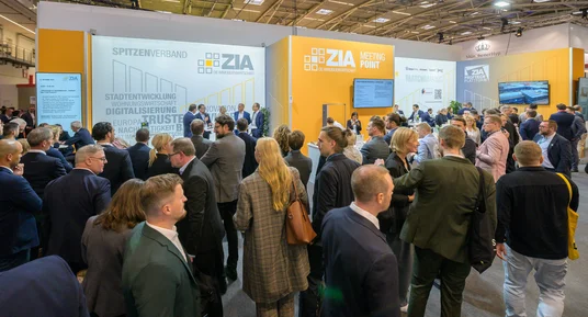 Fotografie einer Messehalle mit vielen Besuchern, die sich an einem großen, orange-weißen Messestand des ZIA (Zentraler Immobilien Ausschuss) versammeln. Stand zeigt Themen wie Stadtentwicklung, Digitalisierung und Nachhaltigkeit, mit mehreren Bildschirmen und Informationswänden, die als Treffpunkt für Networking und Informationsaustausch dienen.