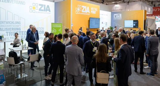 Fotografische Aufnahme einer Messe mit zahlreichen Besuchern am Stand von ZIA, einem Spitzenverband der Immobilienwirtschaft, bei der EXPO Real 2025. Der Stand ist hell gestaltet mit weißen und orangen Flächen, Displays und Texten zu Themen wie Stadtentwicklung, Digitalisierung und Nachhaltigkeit, während sich Fachleute austauschen und Netzwerken.