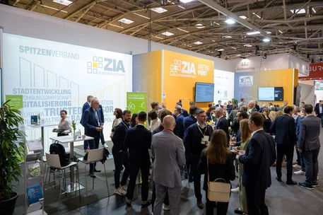 Fotografische Aufnahme einer Messe mit zahlreichen Besuchern am Stand von ZIA, einem Spitzenverband der Immobilienwirtschaft, bei der EXPO Real 2025. Der Stand ist hell gestaltet mit weißen und orangen Flächen, Displays und Texten zu Themen wie Stadtentwicklung, Digitalisierung und Nachhaltigkeit, während sich Fachleute austauschen und Netzwerken.