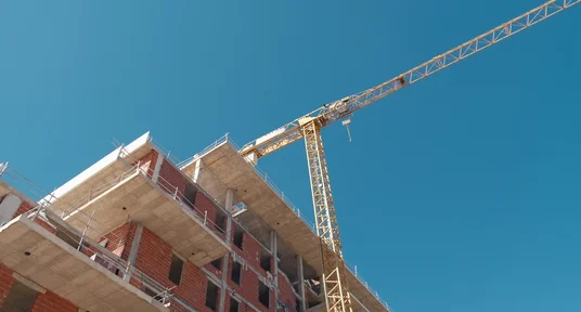 Fotografie einer mehrstöckigen Baustelle mit rohem Ziegelmauerwerk, Betondecken, Gerüsten und gelbem Turmdrehkran vor klarem blauem Himmel. Sichtbare Baufortschritte an Wohngebäude sowie Kranmast und weit ausladender Ausleger verdeutlichen laufende Hochbauarbeiten.