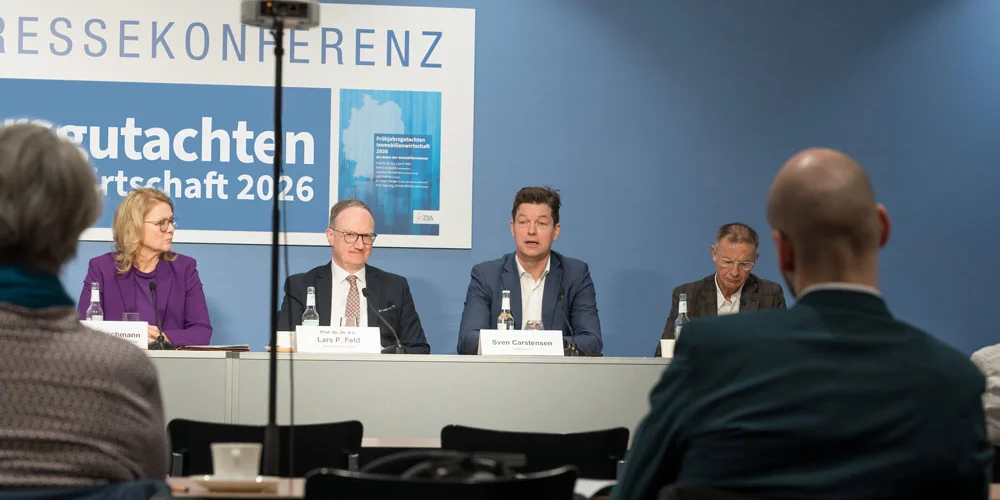 Fotografie einer Pressekonferenz mit vier Fachleuten an einem Podium vor blauem Hintergrund und Präsentationswand „Gutachten Wirtschaft 2026“. Im Vordergrund sitzen Zuhörende, mittig steht eine Kamera; Namensschilder und Dokumente kennzeichnen die Vortragenden.