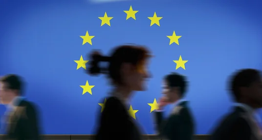Fotografie von vorbeilaufenden, unscharf dargestellten Pendlern vor blauem Hintergrund mit zwölf gelben EU-Sternen. Bewegungsunschärfe und unterschiedliche Silhouetten veranschaulichen Mobilität im europäischen Kontext.