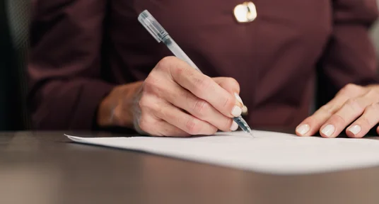 Fotografie einer Person, die mit einem Kugelschreiber ein Dokument auf einem Schreibtisch unterschreibt. Dunkle Kleidung, silberner Stift, weißes Papier und ruhiger Bürohintergrund betonen den formellen Schreibvorgang.