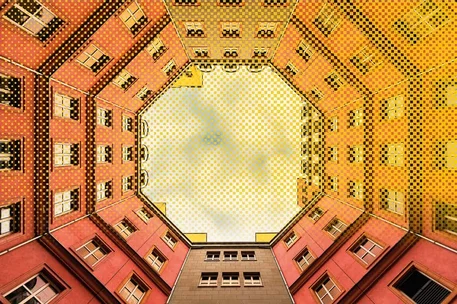 Fotografie eines begrünten Innenhofs mit Blick nach oben, umgeben von roten und gelben mehrstöckigen Gebäudefassaden. Oktogonaler Himmel bildet Zentrum, zahlreiche Fenster, Dachdetails und kräftige Farbtöne betonen symmetrische Architektur und ungewöhnliche Perspektive.