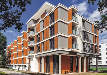 Fotografie eines modernen mehrstöckigen Wohngebäudes mit markanter Architektur und gestaffelten Balkonen. Orangefarbene Klinkerfassade, weiße Betonrahmen, große Fenster, begrünte Außenflächen und blauer Himmel prägen die Ansicht.