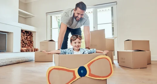 Vater und Sohn zuhause mit Pappflugzeug beim Spielen in einem neuen Zuhause. Freundliche, helle Atmosphäre mit einigen Umzugskartons im Hintergrund.