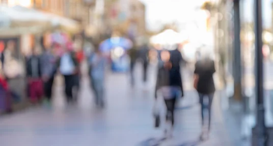 Fotografie einer belebten Fußgängerzone mit unscharf dargestellten Passanten, Geschäften und Marktständen. Helles Gegenlicht, warme Farbtöne und geringe Schärfentiefe lenken den Fokus auf urbane Bewegung und Atmosphäre.
