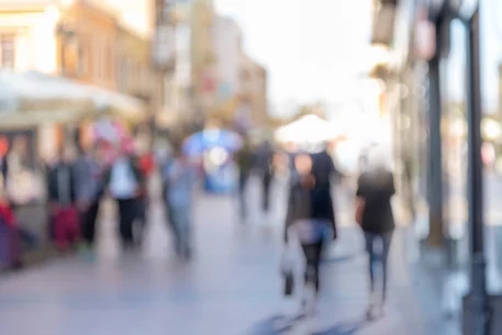 Fotografie einer belebten Fußgängerzone mit unscharf dargestellten Passanten, Geschäften und Marktständen. Helles Gegenlicht, warme Farbtöne und geringe Schärfentiefe lenken den Fokus auf urbane Bewegung und Atmosphäre.