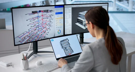 Fotografie eines BIM-Arbeitsplatzes zur digitalen Planung und Koordination eines Gebäudes mit 3D-Modellen. Zwei Monitore zeigen detaillierte Gebäudeansichten mit rot-blauen Leitungsverläufen und Bedienfeldern, während ein Laptop eine weitere technische Draufsicht darstellt.